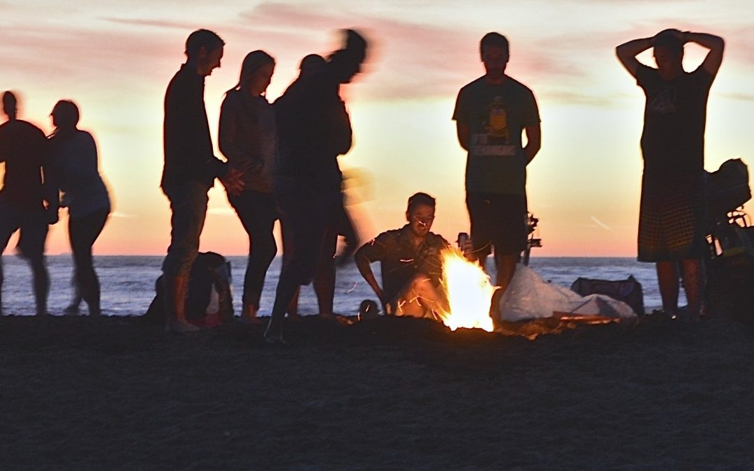 A group of friends having fun with a fire pit BBQ on the beach as the sun sets over the sea