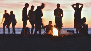 A group of friends having fun with a fire pit BBQ on the beach as the sun sets over the sea