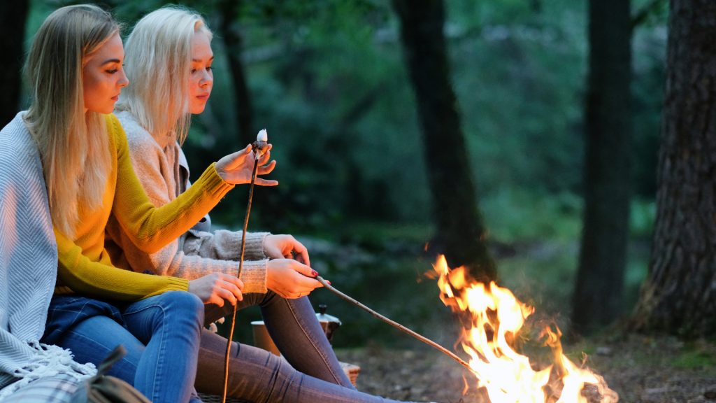 Girlfriends toasting marshmallows together over a fire pit BBQ in a woodland clearing at dusk