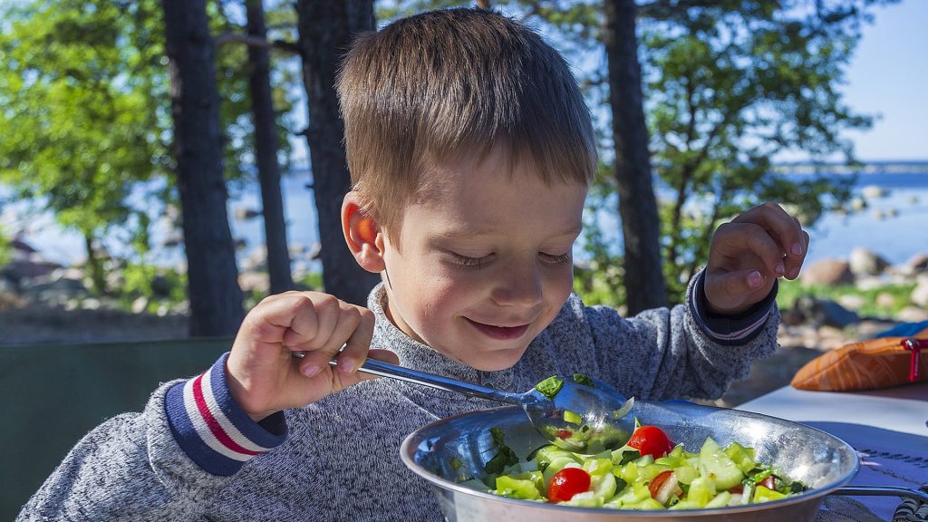 A happy young boy makes salad at a fire pit BBQ party outdoors by a lake