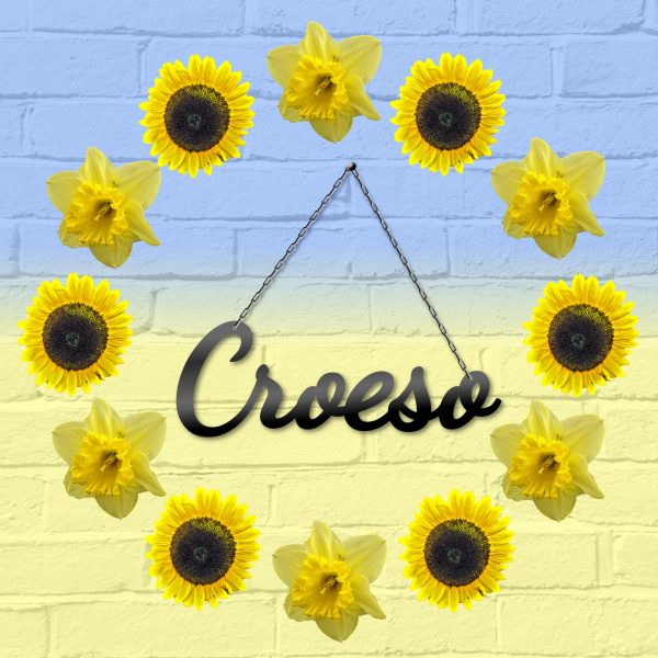 Croeso hanging black metal decorative Welcome in Welsh sign surrounded by Welsh daffodils and Ukraine sunflowers against a wall painted with the Ukrainian flag. This is Ravens' Way Metalworks' Ukraine crisis fundraiser. We will donate 100% of all profits and labour costs from this product to Ukraine Red Cross.