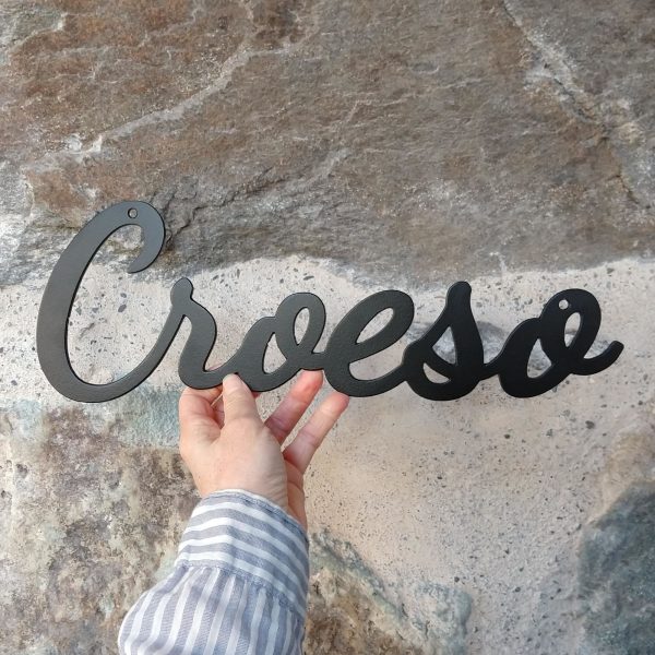 Croeso hanging black metal decorative Welcome in Welsh sign on a white brick wall. This is Ravens' Way Metalworks' Ukraine crisis fundraiser. We will donate 100% of all profits and labour costs from this product to Ukraine Red Cross.