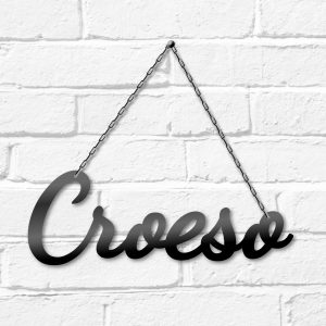 Croeso hanging black metal decorative Welcome in Welsh sign on a white brick wall. This is Ravens' Way Metalworks' Ukraine crisis fundraiser. We will donate 100% of all profits and labour costs from this product to Ukraine Red Cross during March 2022.